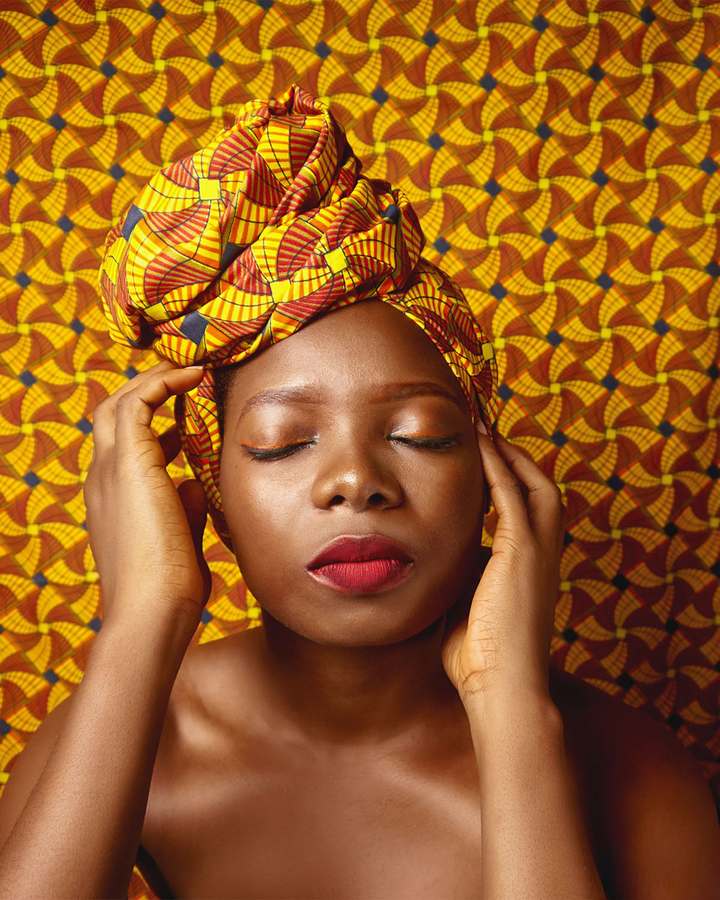 Portrait d'une femme aux yeux fermés, coiffée d'un turban en wax jaune devant un tissu au même motif
